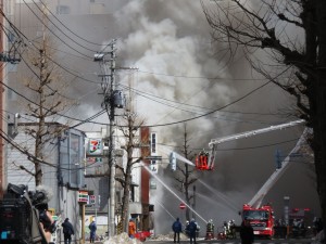 燃えて煙を吐き出している米山商店の建物と消化作業をしている消防隊員さん達。